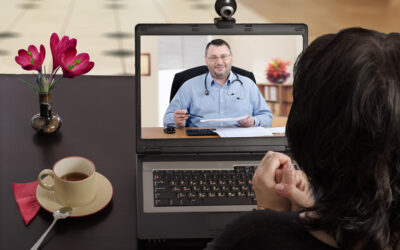 Blackhaired woman sits at a cafe table and consults telemedicine doctor by laptop computer with her back to camera. In monitor, male physician reviews medical laboratory results with her. Horizontal shot on indoors blurred background